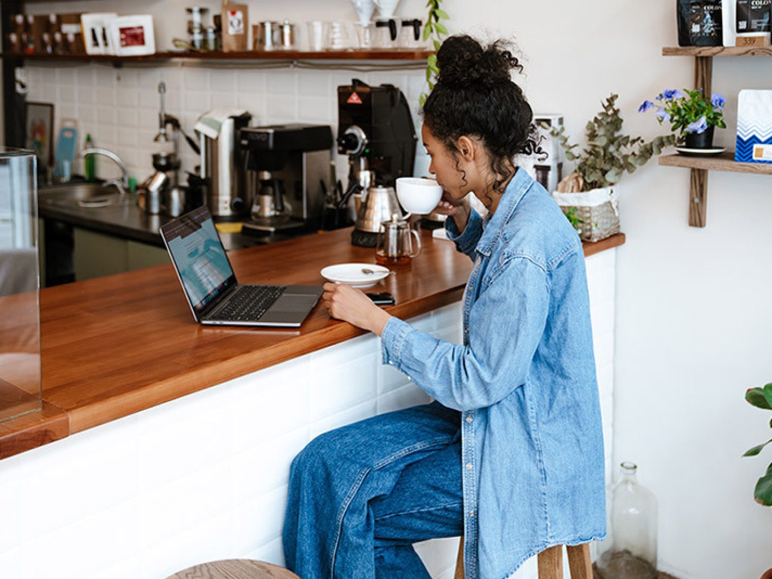 A woman sitting at a coffee bar drinking her cup of coffee and looking at her laptop.