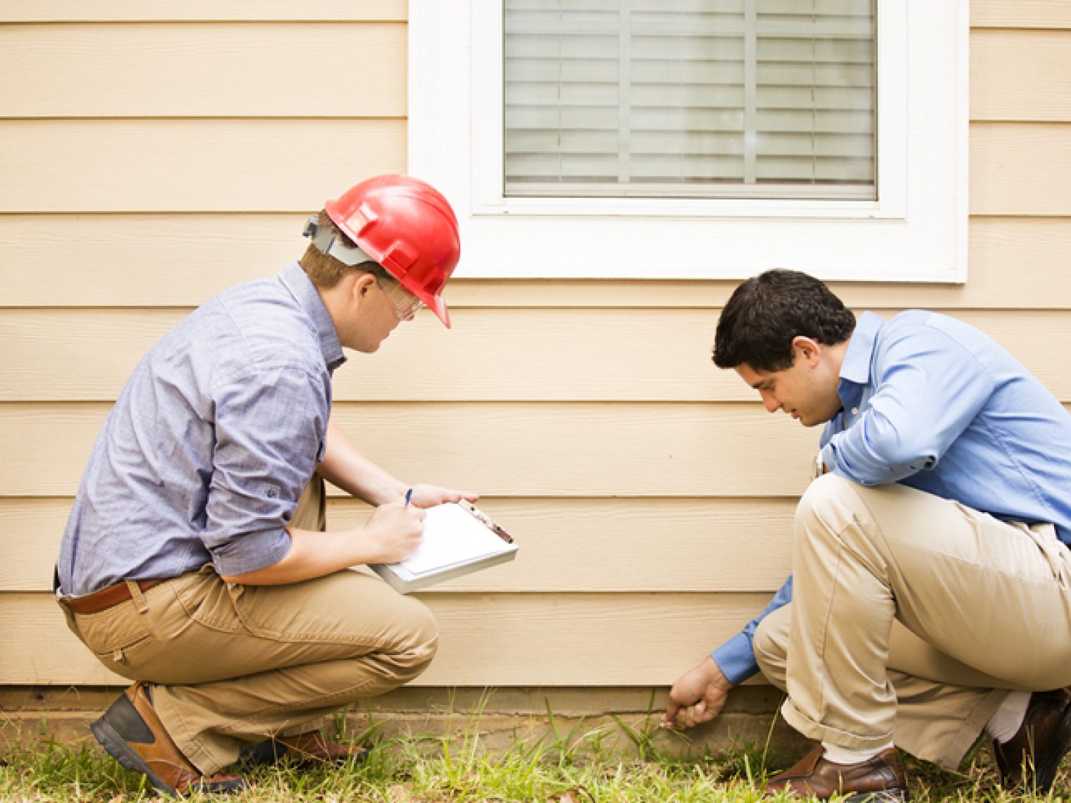 Two men are examining the exterior of a house, with one wearing a red hard hat suggesting that he may be a construction worker, inspector, or engineer. The man with the hard hat is crouched down, taking notes on a clipboard, while the other man is closely inspecting something near the ground.