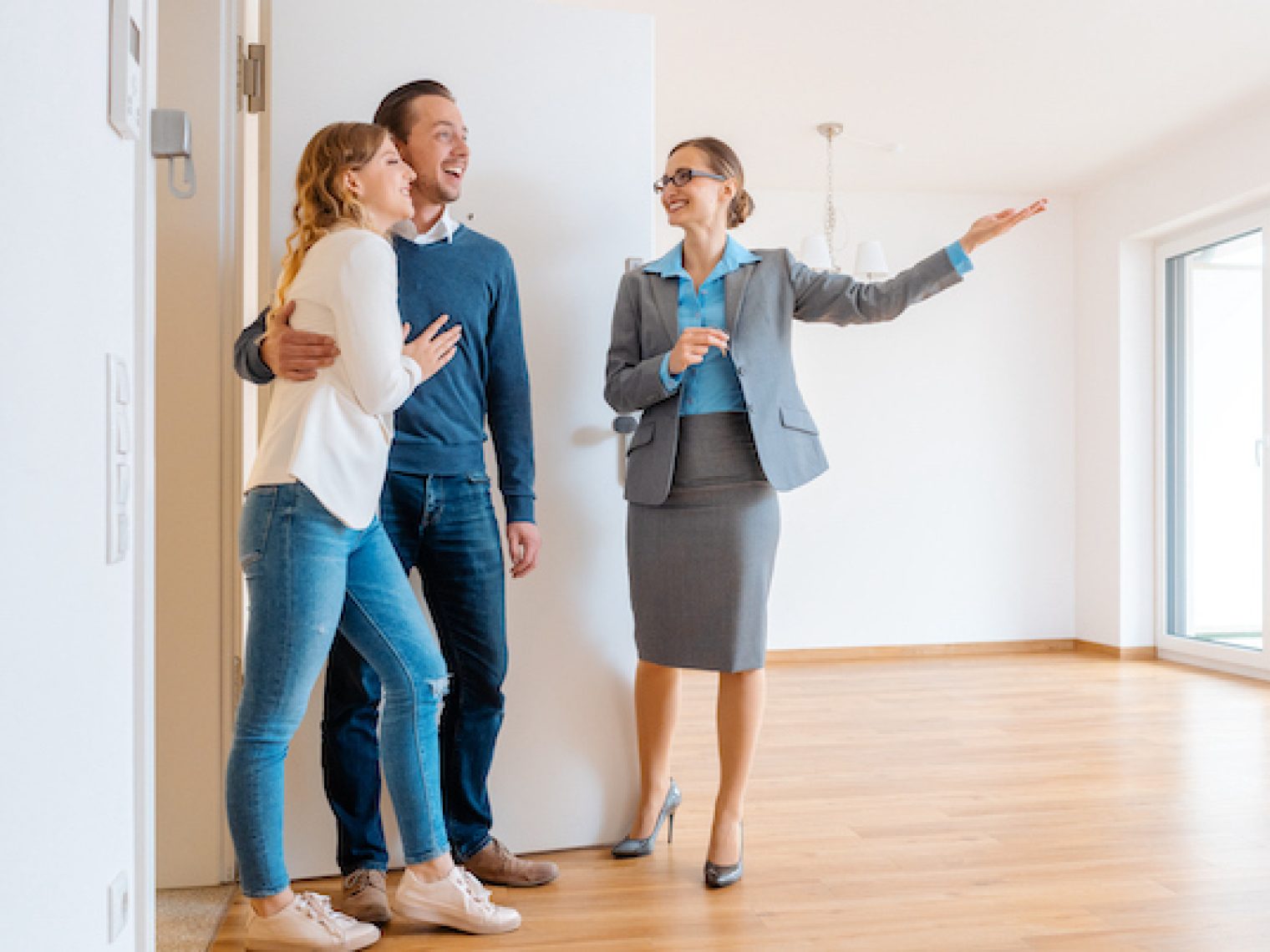 Real estate agent showing a spacious room to a happy couple in a house tour, with the agent gesturing towards large windows with natural light.