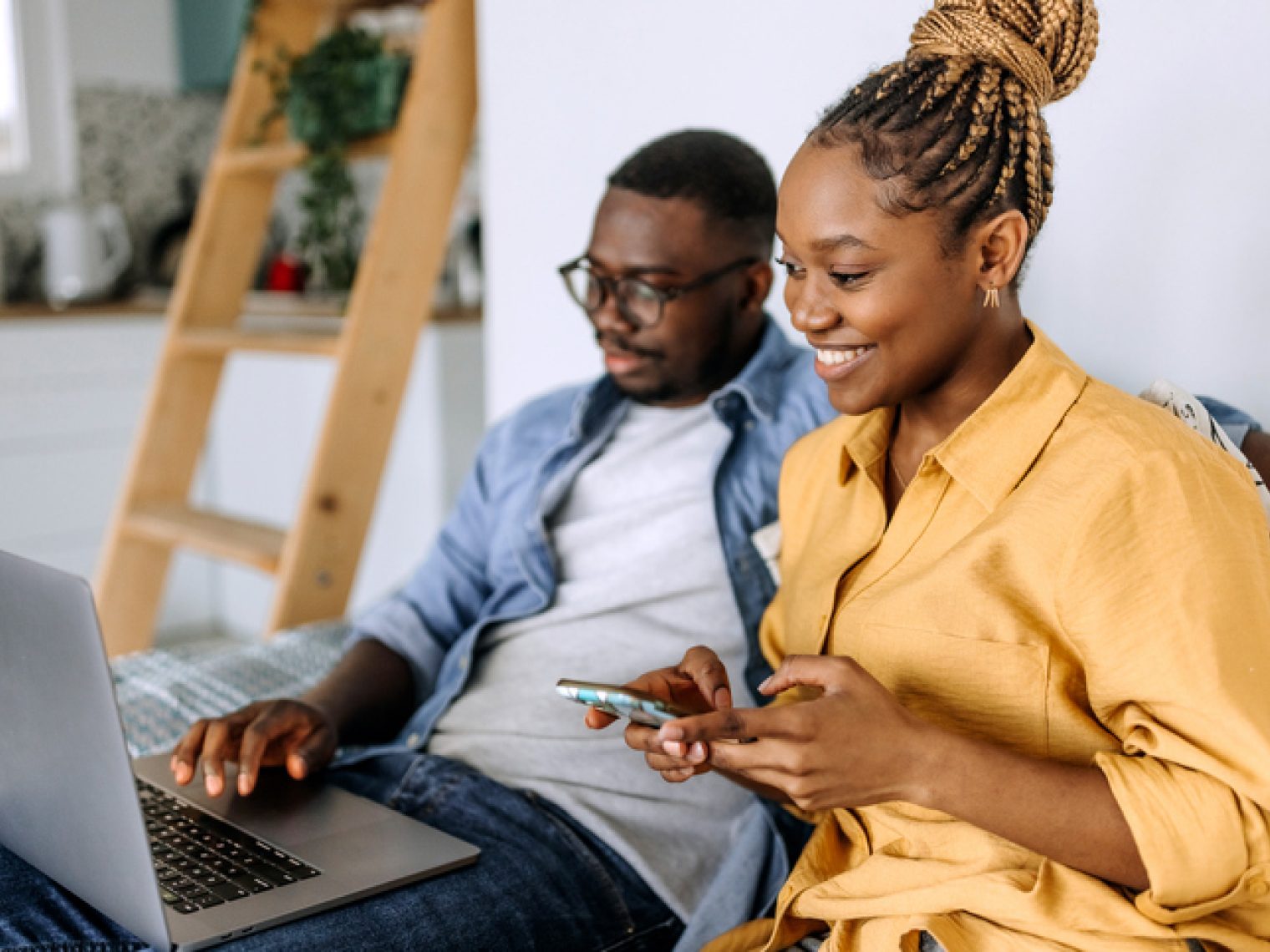 A woman is sitting next to a man who is focused on a laptop. Both are comfortably seated on a couch with pillows, suggesting a relaxed home environment.