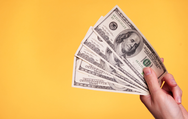 A hand holding a fanned out stack of hundred dollar bills against a bright yellow background.