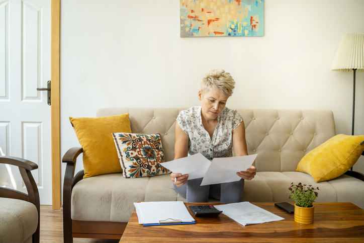 A woman with short blonde hair is sitting on a beige couch, intently reviewing two papers in her hands. She has a focused expression, suggesting she is concentrating on the content of the documents.