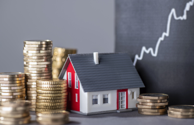 A small red and white model house is surrounded by stacks of coins with a rising chart in the background.