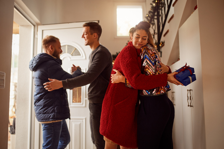 A group of people are engage in friendly greetings at a home entrance. A woman in a vibrant red cardigan and patterned top smiles contentedly as she hugs a man who holds a blue gift box, suggesting a thoughtful gesture or a special occasion.