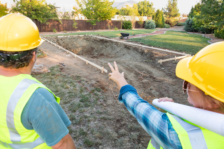 Two construction workers with hard hats and high-visibility vests are discussing over an excavated site for a home addition, with one pointing and holding plans.