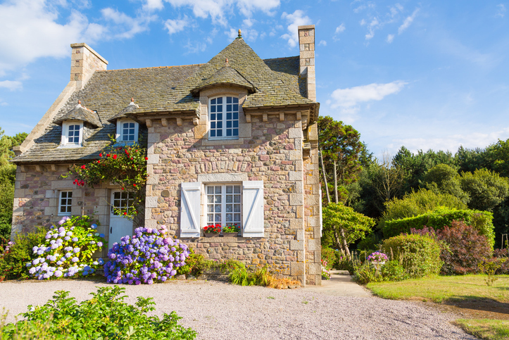 A charming stone cottage with a steep roof, adorned with colorful flowers and shutters, set in a lush garden under a blue sky.