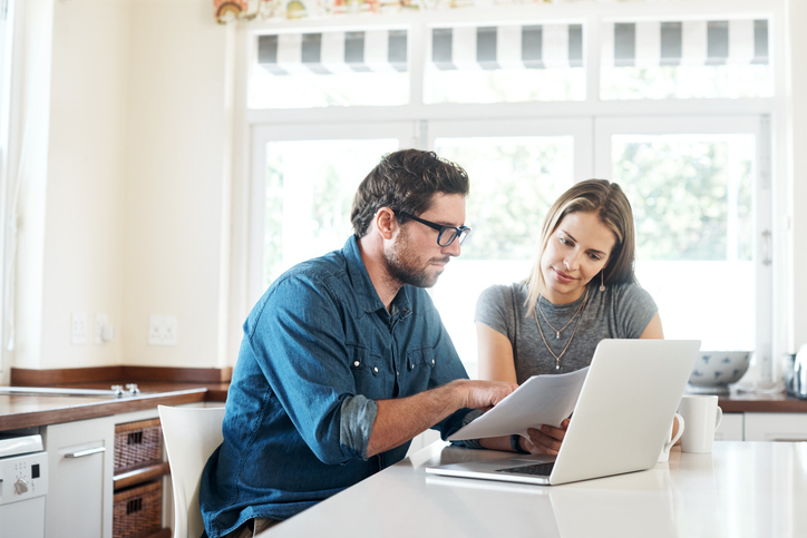 A couple sits at a kitchen table, focusing on documents and a laptop, possibly discussing home warranties or finances.