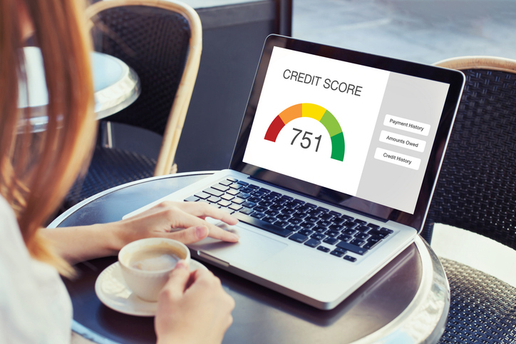 A person is sitting at an outdoor cafe table viewing a laptop screen that displays a credit score of 751, along with credit score components like payment history, amounts owed, and credit history.