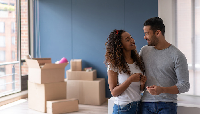 A smiling couple stands close together in a room with unpacked moving boxes, looking affectionately at each other, indicating a new chapter in their lives as homeowners.