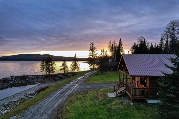 A serene lakeside cabin at dusk with the setting sun casting a warm glow over the tranquil waters and surrounding landscape.