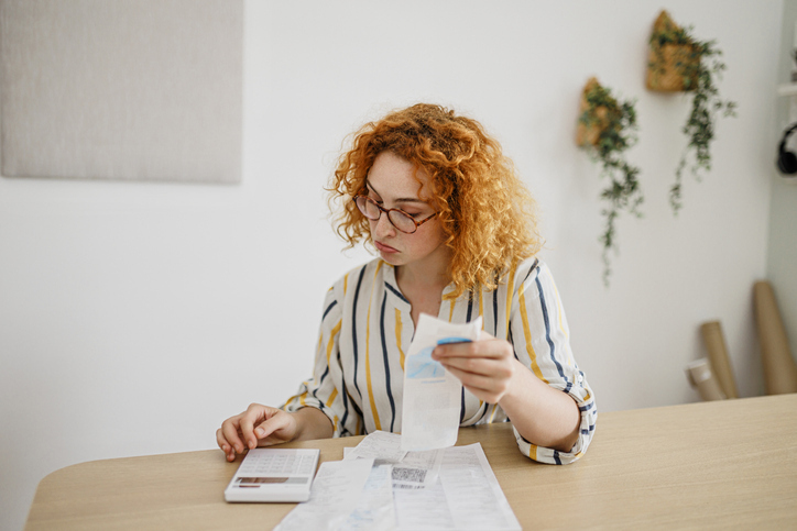 A focused young woman with curly hair and glasses examines receipts and documents spread out on a table.