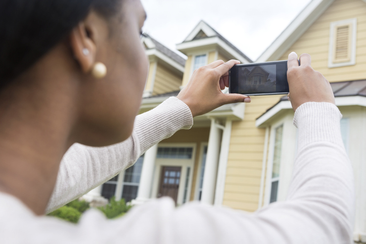 A woman is capturing a photo of a house with her smartphone, potentially for a real estate listing or home inventory.