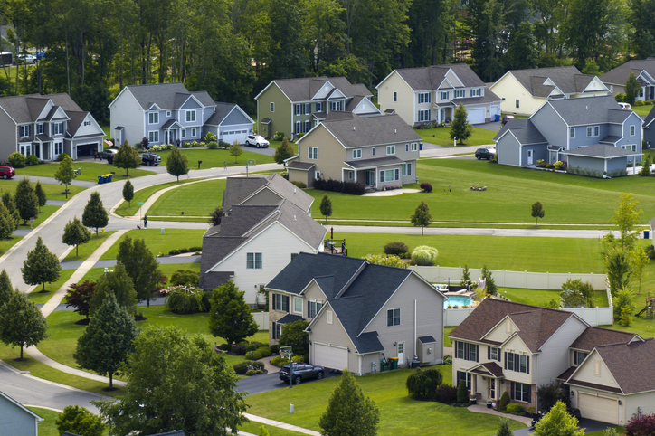 An aerial view of a suburban neighborhood shows a collection of two-story houses, each with its own well-manicured lawn. The homes are architecturally similar, with variations in color schemes and details, presenting a cohesive yet individualized community aesthetic.