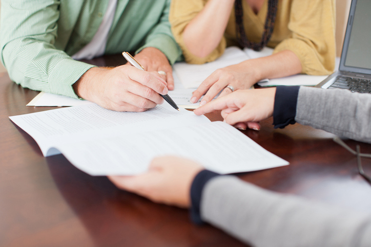 A close-up of three people's hands, with one person pointing at a document while another person writes, with a laptop open beside them on a wooden table.