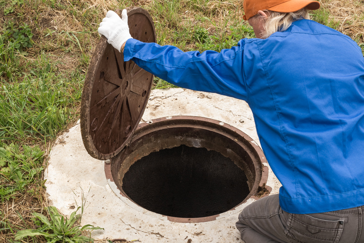 A worker wearing gloves and a blue jacket is opening a large, round sewer manhole cover on a concrete surface with grass around it.