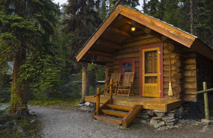 Nestled in a lush coniferous forest, this charming log cabin invites tranquility with its rustic design and natural surroundings. The cabin features a small porch with wooden steps leading to an orange door, complemented by two matching chairs that offer a spot to enjoy the serene environment.