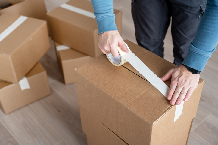 A person is taping a cardboard box shut, part of a stack of boxes, indicating packing or moving activities.