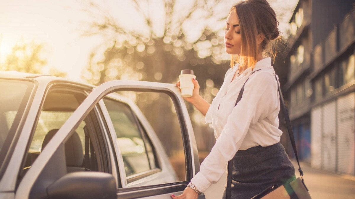 Woman holding coffee cup opens car door.